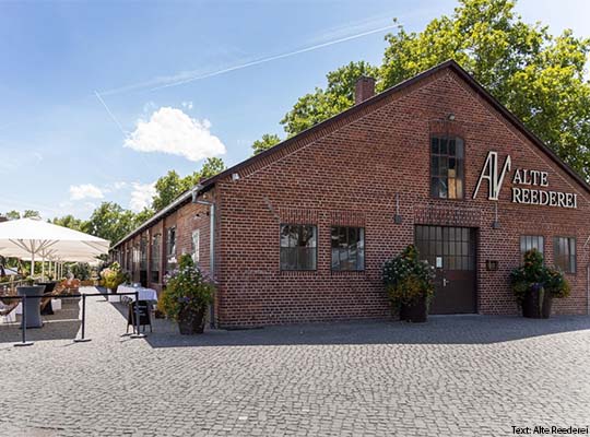 Außengelände der Alte Reederei, im Außenbereich befinden sich kleine Tische auf welchem weiße Tischdecken ausgelegt sind. Das Gebäude ist ein Rotes Backstein-Haus.