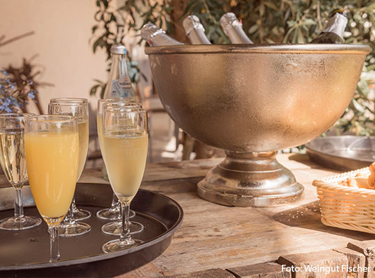 Hölzerner Tisch, auf welchem ein Behälter mit drei Sekt-Flaschen steht, neben dem Behälter stehen drei Sektgläser